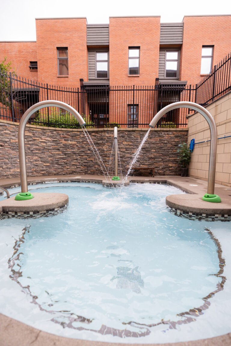 A small communal pool with jets shooting water upward, surrounded by a brick building and black iron fence behind it.